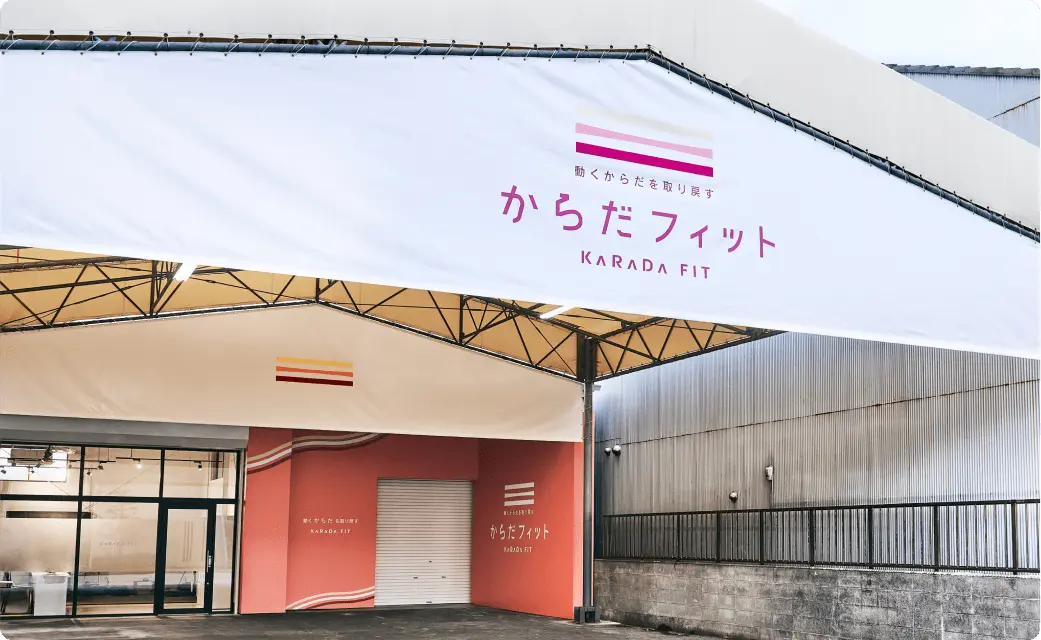 からだフィットの看板がある白い屋根と、ガラス扉・コーラル色の壁が見える建物の外観